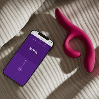 En We-Vibe Nova 2 Rabbitvibrator med en lila "NOVA"-app och en rosa, flexibel klitorisstimulator vilar på en randig tygyta.