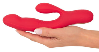 En person håller i en knallröd Thumping Rabbitvibrator, dubbeländad med två böjda silikonförlängningar. Set mot ljus hud, dess eleganta design och "bultande funktion" lovar en förbättrad upplevelse.