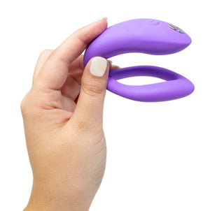 En hand håller WE-Vibe Sync O Vibrator, en lila, böjd silikonenhet för G-punkt-stimulans. Dess eleganta design har en knapp och metalldetaljer på ena sidan, mot en vanlig vit bakgrund.