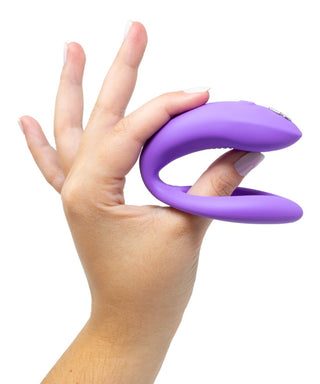 En hand håller en WE-Vibe Sync O Vibrator, en lila, U-formad massageapparat för G-punkt-stimulans. Fingrarna vilar i dess öppna kurva, vilket visar upp dess flexibilitet. Dess släta struktur sticker ut mot den vita bakgrunden och framhäver dess eleganta innovation.