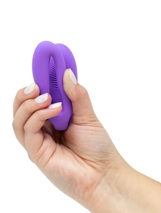 En hand håller i en WE-Vibe Sync O Vibrator, en liten lila silikonenhet med en öppning i mitten, som liknar en elegant teknisk pryl eller livsstilstillbehör. Naglarna är målade vita mot en vanlig vit bakgrund.