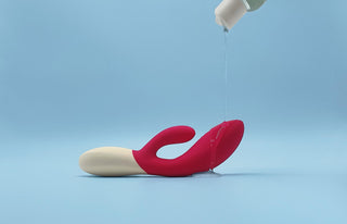En röd och vit rabbitvibrator ligger på ett blått underlag, täckt med glidmedel som har hällts över den. Produkten är centrerad i bild mot en enkel blå bakgrund.