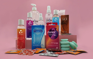 En samling intimprodukter uppställda mot en rosa bakgrund. Bilden visar glidmedel från Easyglide och Just Glide, kondomer från Durex och Pasante, en brun förpackning från Slow Sex, samt flera små färgglada förpackningar och gröna penisringar i silikon