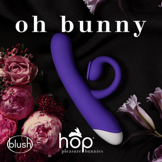 HOP - Oh Bunny - Sugande Rabbitvibrator (19 cm) sitter elegant bland rosa blommor med logotyper från Blush and Hop Pleasure Bunnies på en elegant svart bakgrund. Tillverkad av högkvalitativt silikonmaterial utlovar den en oförglömlig upplevelse med sin integrerade klitorisstimulator i livfull lila.