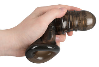 En hand håller ett mörkt, flexibelt, räfflat silikonobjekt med en cirkulär bas som liknar Rebel Dick & Ball Penis Sleeve.