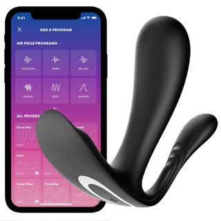En svart, kurvig Satisfyer Top Secret + silikonenhet sitter bredvid en smartphone och visar en app med alternativ som Good Fun, Spark och grafiska vågmönster. Båda föremålen har en elegant, modern design.