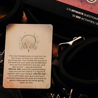 Ett kort från Box Of Burning Desires, med intima instruktioner, placeras bredvid svarta handbojor på en träyta.