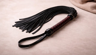 Flogger med svart handtag och mjuka läderremmar placerad på ljus bakgrund, fotograferad i lugn och stilren miljö.