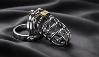 Stilren cock cage i rostfritt stål med lås, fotograferad mot mörkt tyg – exempel på kukbur för kyskhet och kontroll