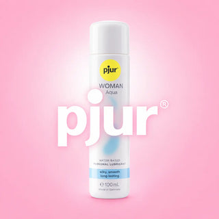 Pjur Woman Aqua vattenbaserat glidmedel 100 ml i vit flaska, mot en mjuk rosa bakgrund med vit Pjur-logotyp.