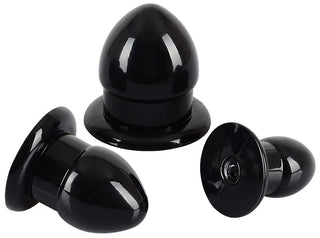 Anal Stretching Plug Kit har tre svarta gummipluggar med kupolformade toppar och platta baser, mot en vit bakgrund. De varierar i storlek, med den största upprätt, en liggande på sidan, och den minsta också upprätt.