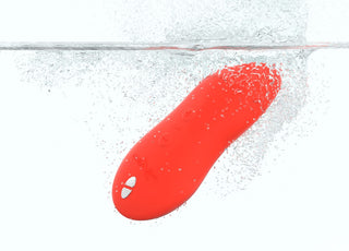 En We-Vibe Touch X-vibrator, i klarrött och helt vattentät, plaskar ner i det klara vattnet medan bubblor bildas runt den eleganta enheten.