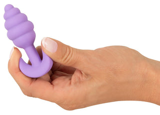 En hand håller i Cuties Mini Buttplug Lila, gjord av kroppssäkert silikon, dess texturerade lila yta kontrasterar mot en vit bakgrund.