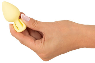 En hand håller en Cuties Mini Buttplug Gul med grepphandtag, tillverkad av kroppssäkert silikon, mot en enkel bakgrund.