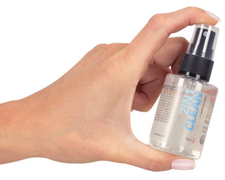En hand håller i en liten genomskinlig sprayflaska märkt "2in1 Clean Intim & Toy Cleaner", perfekt för veganvänlig rengöring av intima föremål och leksaker, mot en vit bakgrund.