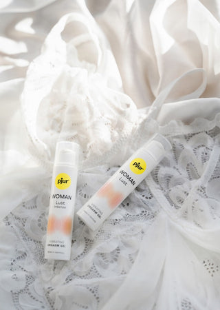 Två flaskor Woman Lust Intense gel vilar på ett delikat vitt spetstyg, som lovar intensiv njutning med naturliga ingredienser.