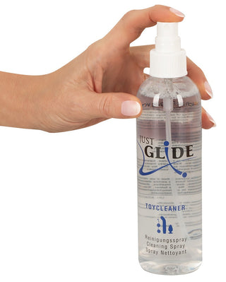 Håller i en klar sprayflaska märkt Just Glide Rengöringsspray 250ml med ett vitt munstycke - en alkoholbaserad formula som är perfekt som rengöringsspray för sexleksaker.