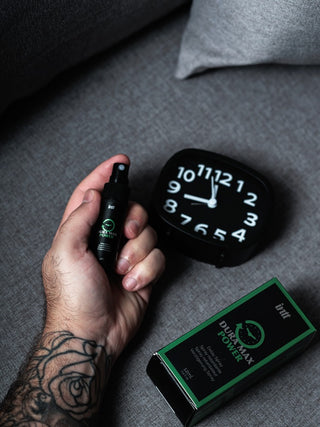 En tatuerad hand håller i Dura Max Power Delay Spray 12ml-flaskan nära en klocka och en grönmärkt låda på en grå yta, och lyfter fram en effektiv spray för män som hjälper till att förlänga njutningen.