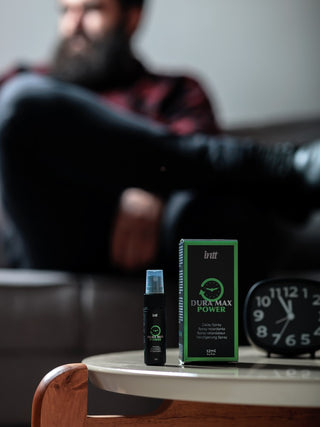 En Dura Max Power Delay Spray 12ml och dess förpackning ligger på ett bord bredvid en klocka, med en suddig man i bakgrunden.