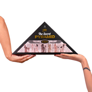 Två händer håller en svart pyramidformad låda märkt Erotiskt Spel Pyramid med egyptisk konst - ett brädspel för vuxna som är perfekt för fans av erotiska spel och nöjen med Egypten-tema.