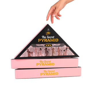 En hand lyfter Erotiskt Spel-pyramiden, en triangulär svart låda, ovanför två rosa rektangulära lådor, vilket antyder ett vuxenspel med Egypten-tema.