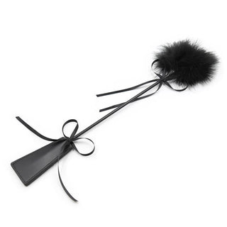 Tickler & Paddle 2-i-1 är en BDSM-leksak i svart läder med en fluffig fjäderpom-pom och bandrosett på handtaget – perfekt för lekfulla bondageäventyr.