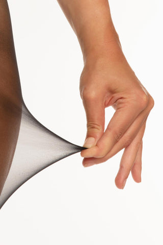 En hand sträcker det skira svarta Christine Tåliga Strumpbyxor-tyget mot en vit bakgrund och visar upp dess genomskinlighet, elasticitet och anti-stegeteknologi för ökad hållbarhet.