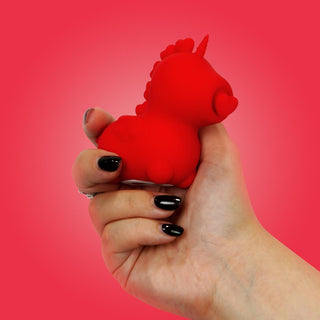 En hand med svart nagellack håller en liten röd enhörningsleksak mot en livfull rosa bakgrund, vilket speglar de ergonomiska kurvorna hos Unihorn Cupids Beau Klitorisvibrator.