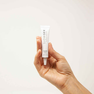 En hand håller en vit tub märkt Slow Sex Klitorisbalm 10 ml, som visar detta sensuella balsam mot en enkel ljus bakgrund.