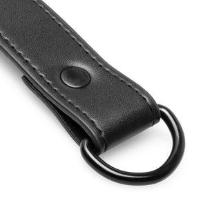 Närbild på Bondage X-Kors Hogties svarta läderrem med sömmar, en metallnit och en rundad D-ring, utformad för användning vid fasthållningslekar.