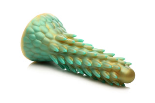 Stegosaurus Dildo Silikon, med en grön och guld drakskalig textur, som påminner om en dinosauriedildo, vilar på en vit yta.