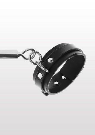 Taboom Bondage Spreader Bar med ankelmanschetter, med svarta lädermanschetter med silvernubbar och en metall D-ring på vänster manschett, är inställd mot en gradient bakgrund som övergår från ljus till mörkgrå.