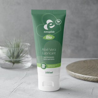 En grön tub med Aloe Vera Bio Glidmedel 100ml står på en grå yta, med naturliga ingredienser och en liten växt i bakgrunden.