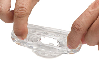 Två händer sträcker ut Transparent Ball Stretcher XR, en klar, flexibel ring med en strukturerad inre yta och små ojämnheter, mot en vit bakgrund.