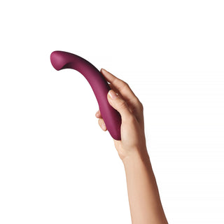 En hand håller Dame Arc G-punktsvibrator - en böjd, rödbrun vibrator avsedd för klitorisstimulering - mot en vit bakgrund.