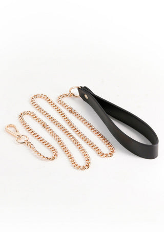Fetish & Fashions Nyx Koppel är ett lyxigt guldkedjekoppel med ett svart läderhandtag, perfekt för ett elegant tillskott till din bondage-kollektion, allt presenterat på en orörd vit bakgrund.