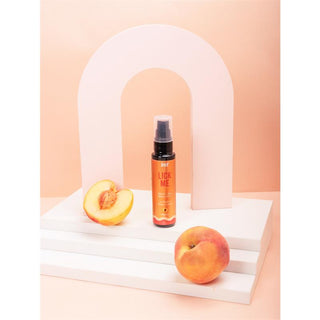 En flaska Intt Lick Me Persika 50 ml sitter lockande på en vit plattform, omgiven av läckra persikor och mot bakgrund av en rosa båge, och lovar sensuella upplevelser.
