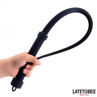 En hand håller i Latetobed Silicone Whip 61 cm med slingrande ände och texturerat handtag; Latetobed-logotyp i hörnet. Perfekt för dominanslekar och för att utforska nya sidor av BDSM.