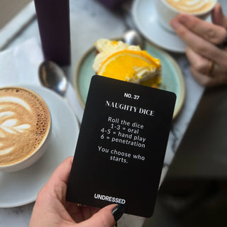 En hand håller en Undressed Intimacy Cards Kortlek för par ovanförför ett bord med kaffe och dessert - perfekt för att stärka relationen med ett roligt och intimt parspel.