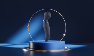 En elegant svart Glam Dual Wand Vibe vilar på en blå piedestal med en guldring, markerad under en spotlight, vilket skapar en lockande fokuspunkt mot den mörka bakgrunden.