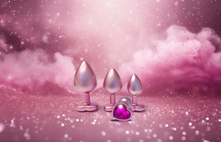 Tre metallicrosa hjärtformade pluggar med ädelstenar vilar på en gnistrande rosa bakgrund fylld med moln och glitter, vilket framhäver charmen hos Pearl Pink Buttplugg Small.