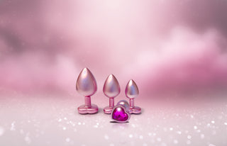 Tre metallicrosa aluminium Buttplugs med hjärtformade baser och en Pearl Pink Buttplugg Medium med en lila hjärtpärla, allt på en glittrande rosa bakgrund.