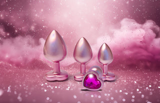 Pearl Pink Buttplugg Large, en metallisk hjärtformad plugg med en gnistrande rosa ädelsten, visas på en glittrande rosa bakgrund med moln.