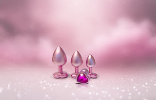 The Gleaming Love Pearl Pink Buttplug Set, med hjärtformade metalliska baser, lyser elegant mot en drömmande rosa grumlig bakgrund och erbjuder både sofistikering och lekfull tjusning.