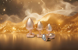 Tre metalliska buttpluggar i aluminium med ädelstenar från vårt Pärlguld Buttplug Set 3-pack lyser på en reflekterande gyllene yta, omgivna av glitter och rök - upplev sann elegans.
