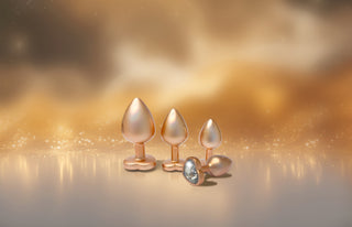 Tre metalliska pärlguld buttpluggar i olika storlekar vilar på en reflekterande yta med en gyllene, suddig bakgrund - ett elegant Pärlguld Buttplug Set 3-pack för alla önskningar.