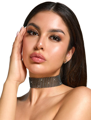 Mot en vanlig bakgrund rör en person med långt mörkt hår ansiktet. De har på sig en Svart Glittrande Choker prydd med strasstenar, kompletterad med glansiga läppar, djärv ögonmakeup och slät hud.