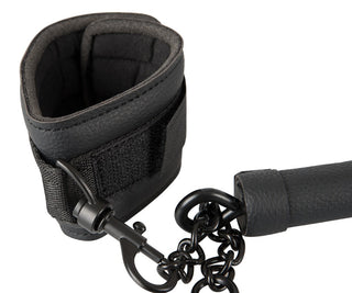 Svart vadderad handledsfixering med kedja och clips, så kallad Spreader Bar med Bojor, mot vit bakgrund.