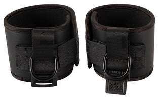 Sida vid sida mot en vit bakgrund fångar två svarta lädermanschetter med kardborrband och D-ringar i metall den eleganta kanten på Badkittys "Handbojor - Spank Me"-kollektion.