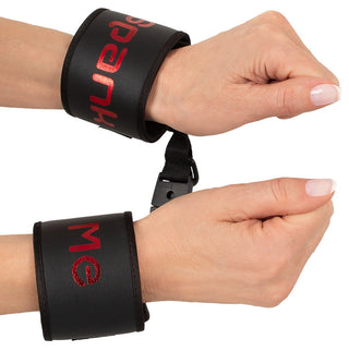 Närbild av händer som bär Badkitty Handbojor - Spank Me svarta handledsmanschetter med röd text, sammankopplade med en liten svart klämma, ekande edgy stil.